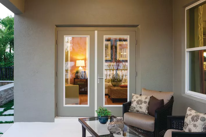 A covered back patio with two chairs and a table connected to the living room of a home by hinged patio doors.