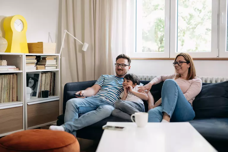 A family sitting on the couch watching TV together in a living room that is illuminated by natural light from their energy-efficient windows.