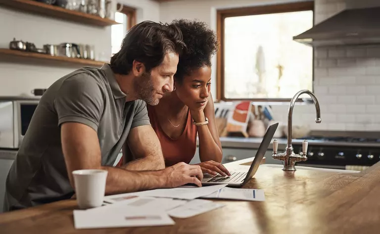 A man and a woman in the kitchen looking at a laptop with papers around it.