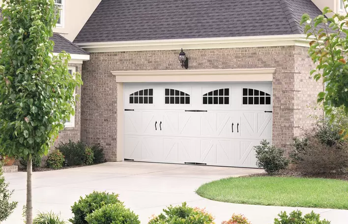 A beautiful, white modern garage door with black handle accents and tan trim on a grayish-brown brick house.