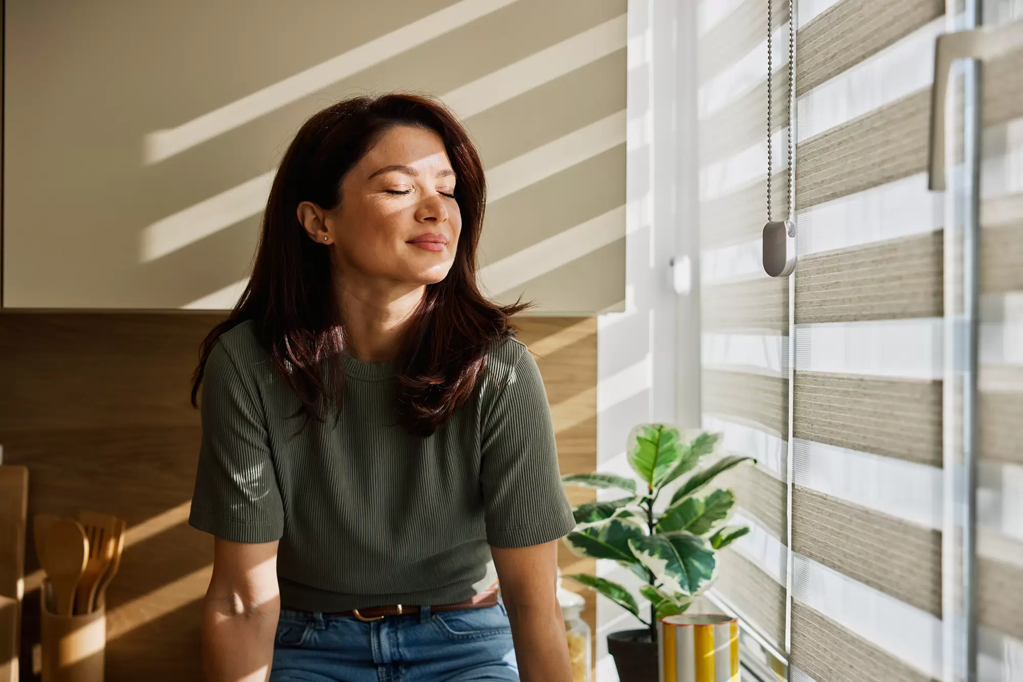 Woman sitting beside a window with sunlight filtering through the blinds.