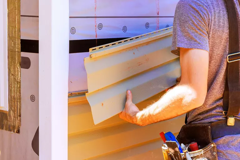 Person stripping away a piece of tan vinyl siding from a home.