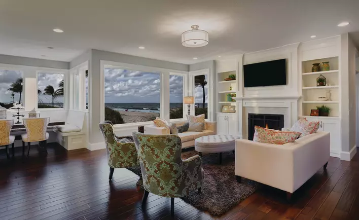 The interior living and dining area of a home with dark hardwood floors, neutral-toned furniture, and walls covered in impact picture windows overlooking the beach.