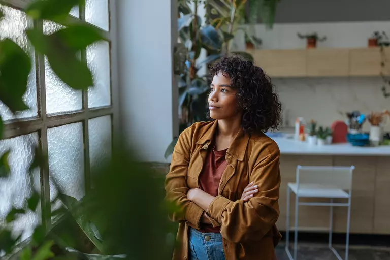 Young woman looking out a window, contemplating in her modern, plant-filled apartment.