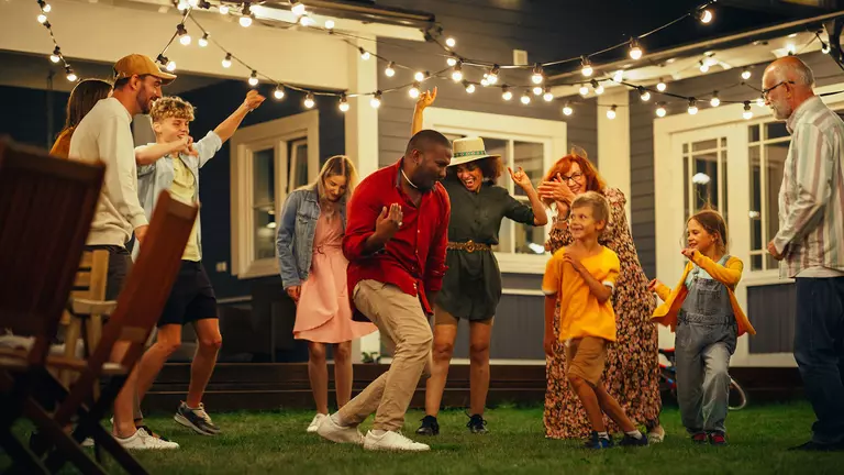 A group of family and friends dancing in their back patio and yard with string lights and modern furniture decorated around them.
