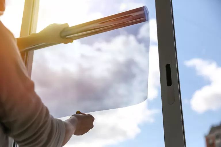 A person applies insulating window film to a casement window on a sunny day to keep the summer heat out.