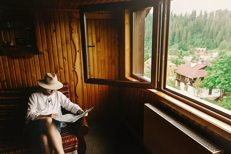 A man reading the newspaper in a nook of his log cabin home, with his casement windows open to let all the fresh Blue Ridge air inside.