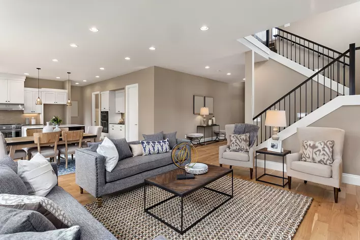 A large, open living room connected to the kitchen and stairs that is decorated with gray
and beige furniture with wooden accents.