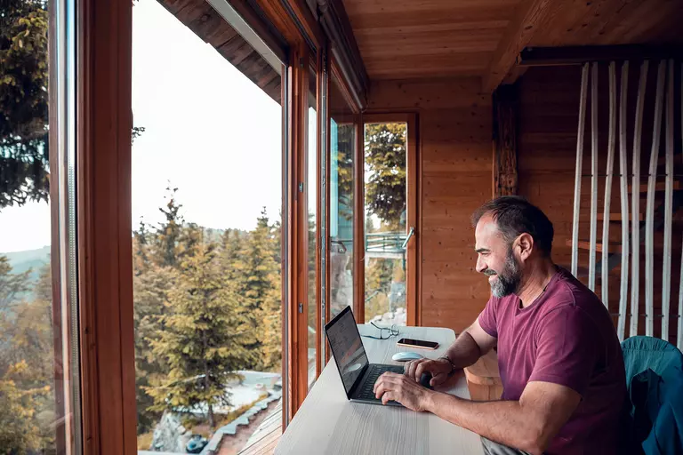 A man working on his laptop in the home office of his log cabin home, looking through large picture windows out into the mountainous landscape beyond.