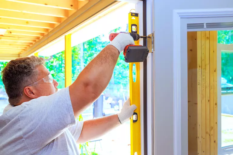 A contractor using a level and a drill while installing a door hinge.