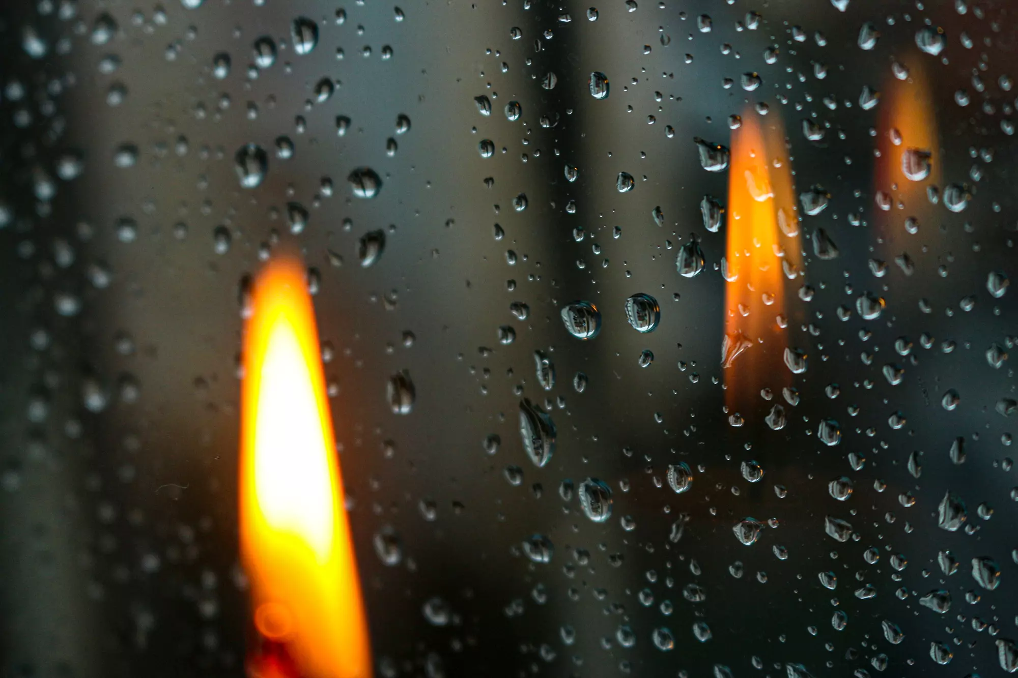 A lit candle in front of a window pane with many water droplets.