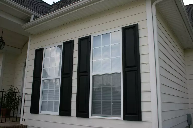 Close-up of windows with black exterior window shutters on the side of a beige-colored home.
