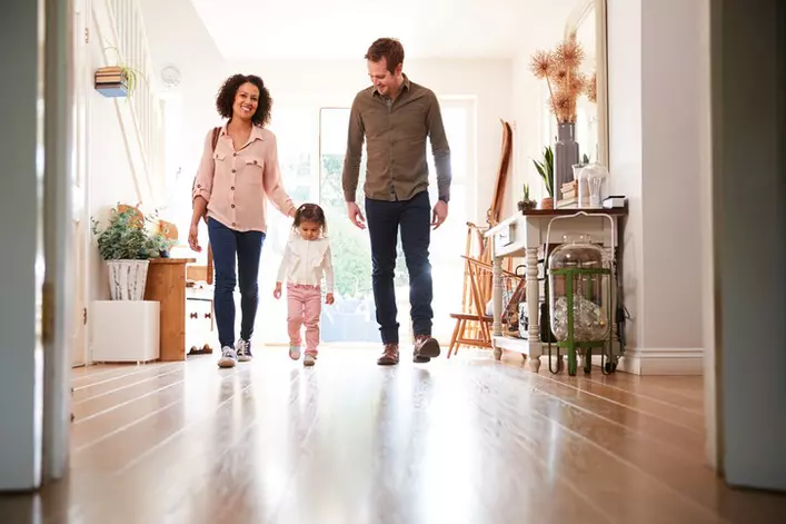 A mother, father, and daughter enter their home and walk through the foyer. They are backlit by natural light coming through their storm door.
