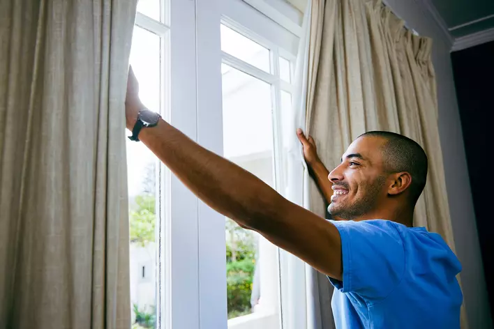 A man smiling and opening his curtains to a beautiful, sunny day outside.