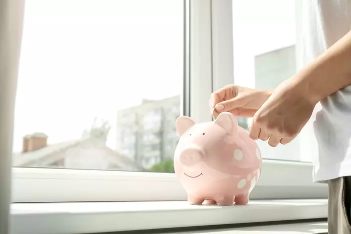A person inserting a coin into a pink piggy bank on the ledge of a vinyl window inside of a home.