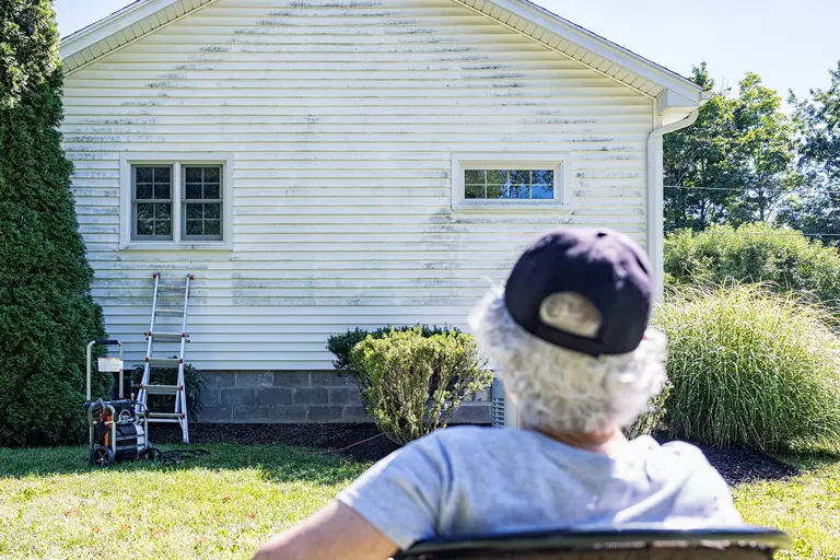 An individual looks at pressure washing equipment in front of a home’s side with black mold growth.