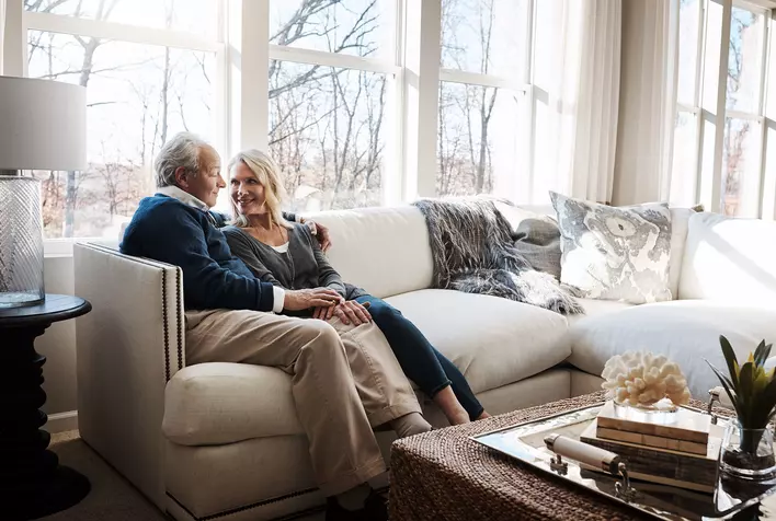 An older married couple sitting on a couch in a living room that is brightly lit by the
natural light of several large windows.