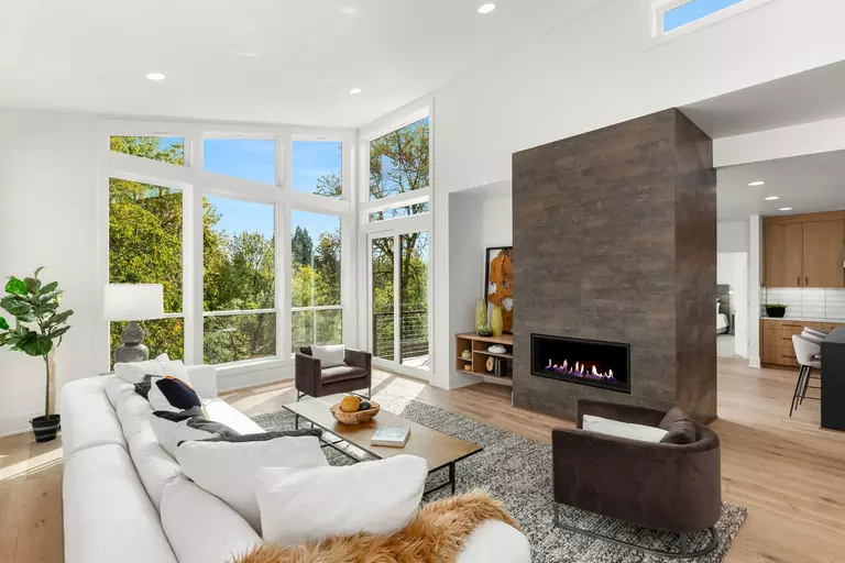 A modern home with a fireplace, a white couch in the living room, and broad windows overlooking a forest.