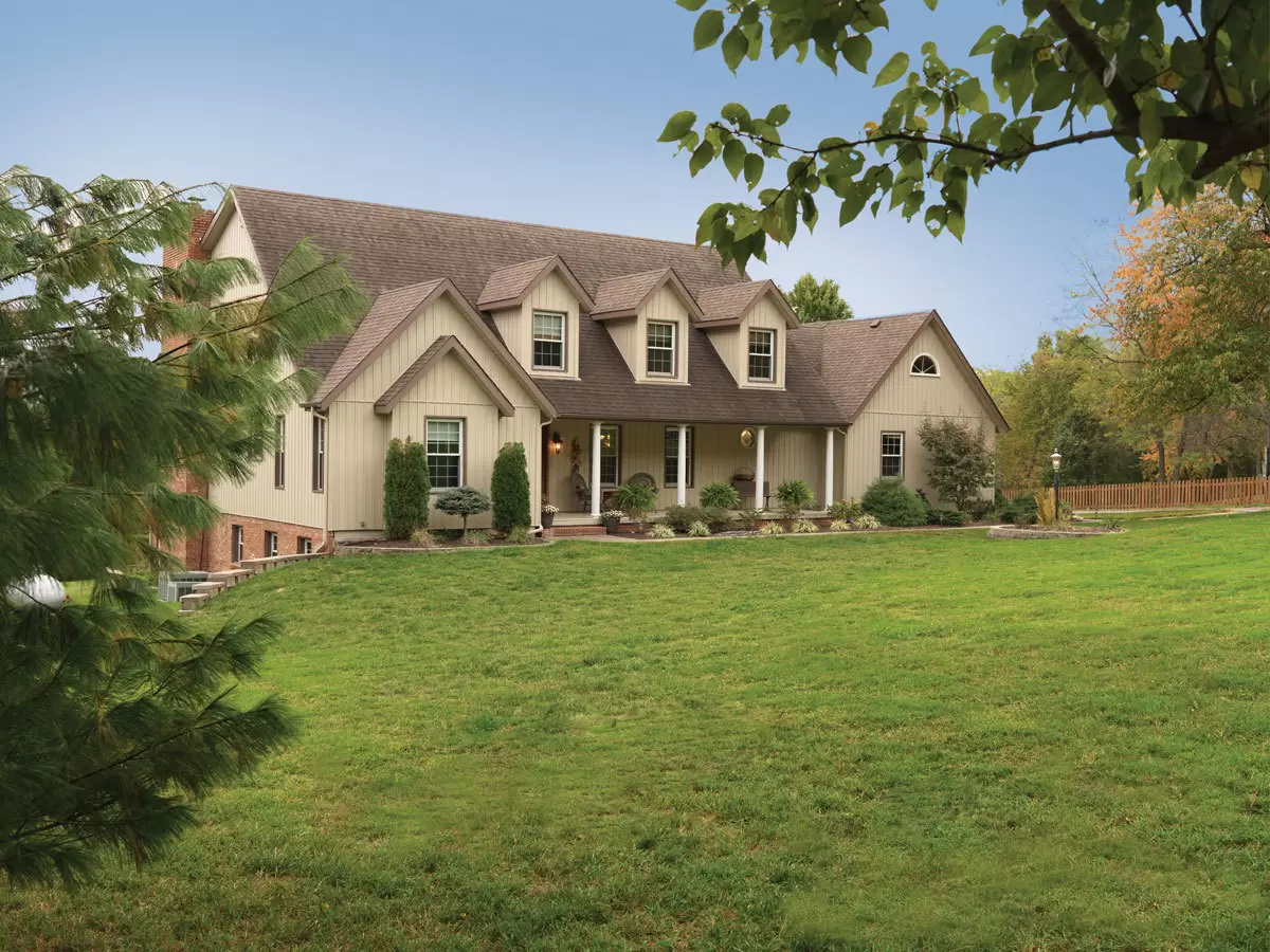 Tan home with board-and-batten siding, dormer windows, and a covered front porch on a large lawn.