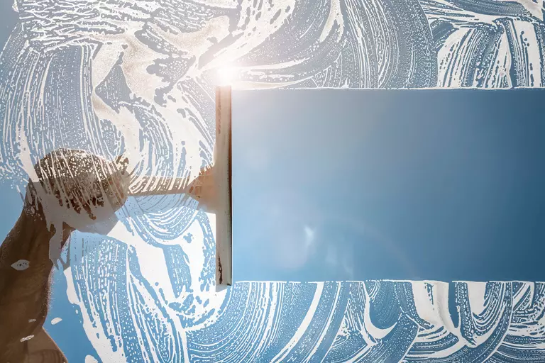 A person moves a squeegee across a pane of glass covered in soapy liquid.