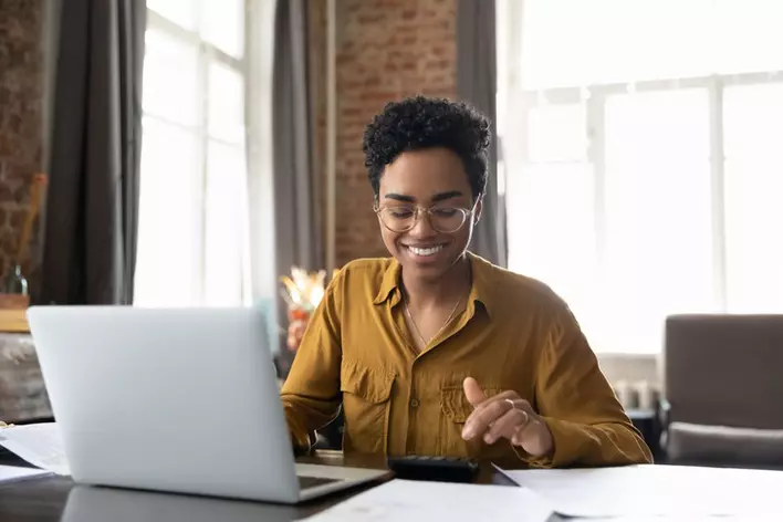Happy young woman in glasses calculating expenses with a calculator and a laptop computer while sitting at a table in her home. 