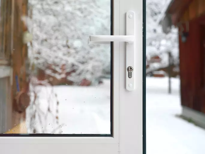 The handle of a white storm door, pictured against a snowy background. The door is opening and has been photographed before swinging shut.