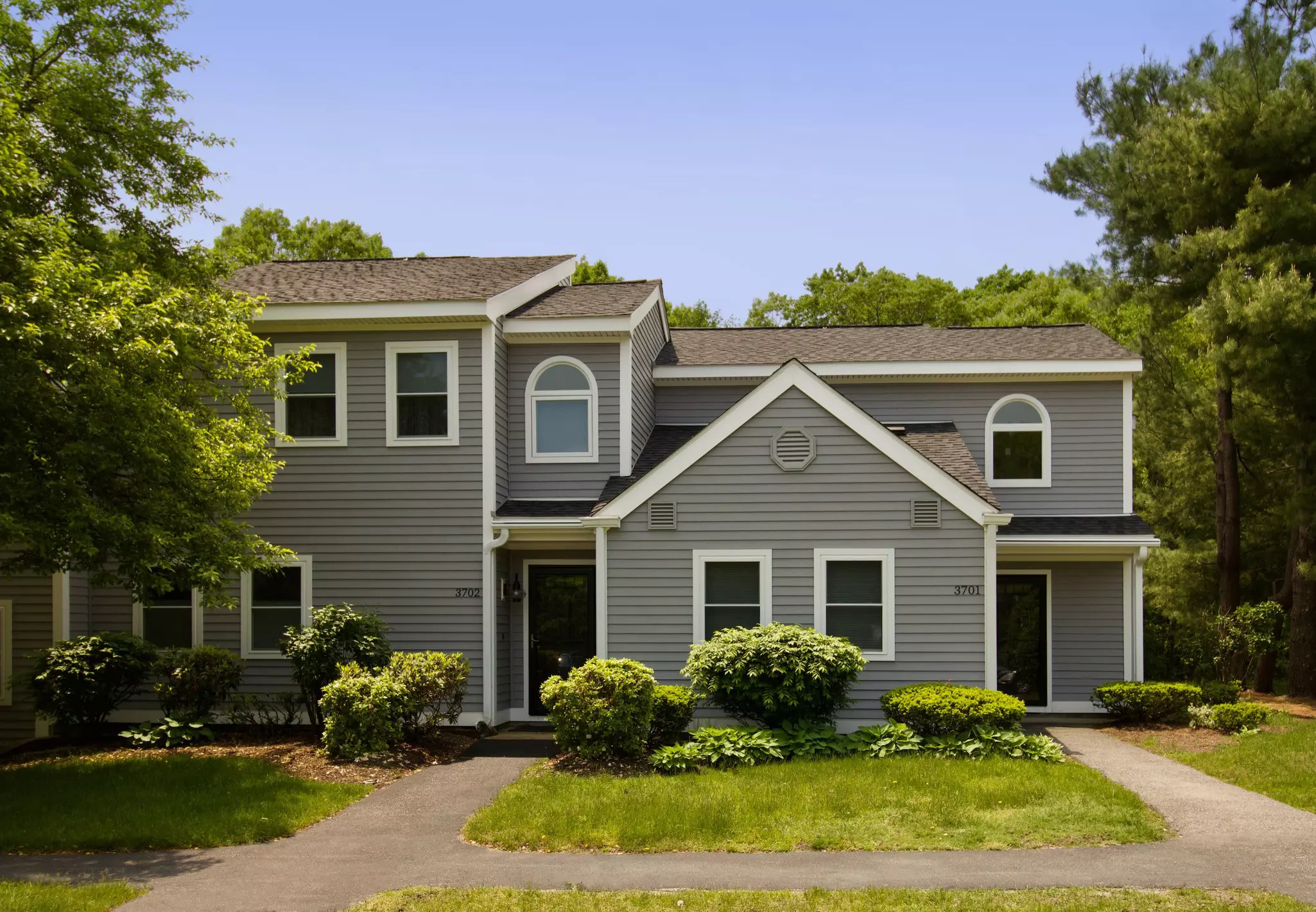 Two-story suburban home with light gray vinyl siding, white trim, and lush green landscaping.