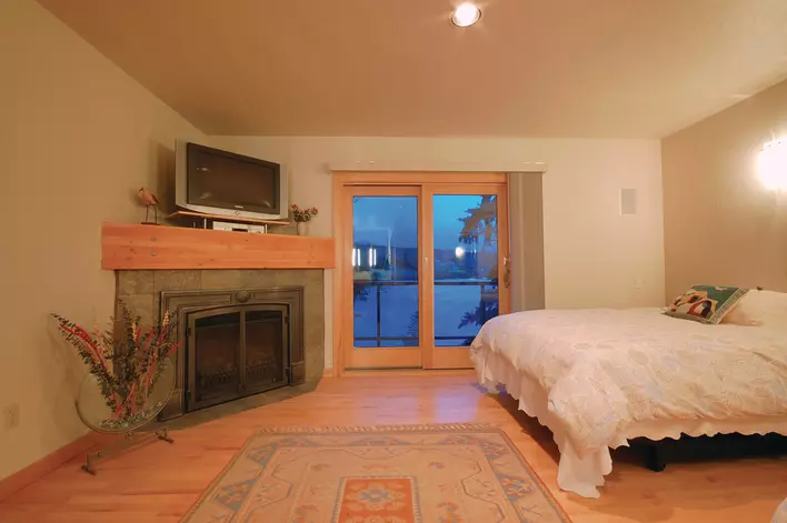 An earth-toned master bedroom with a fireplace, TV, and sliding patio doors leading to a balcony overlooking a lake.