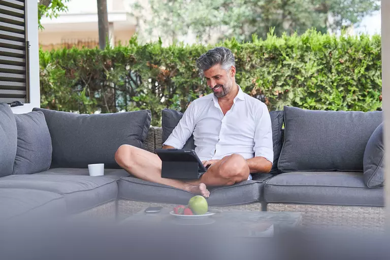 A man types on his laptop while sitting on outdoor furniture in his backyard.