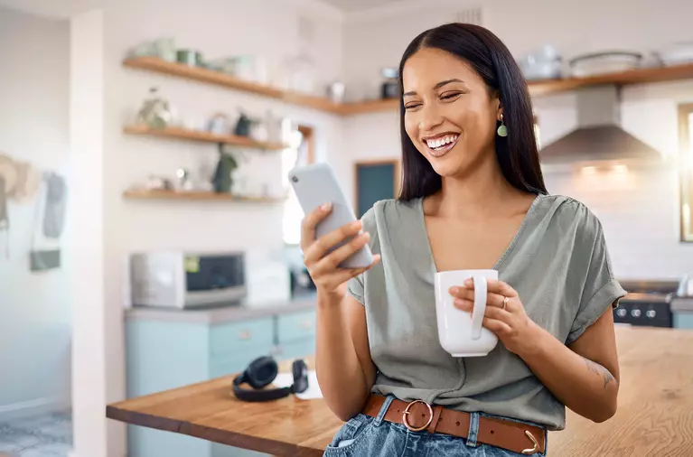 A woman in a kitchen looking at her phone and laughing.