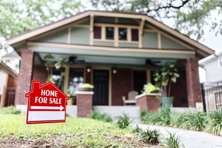 A for-sale sign in front of a home with brick and light gray exterior colors.