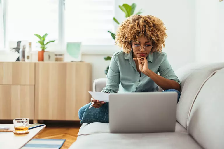 Afro-American woman is sitting on a sofa and looking at a laptop while holding a paper in her hand.