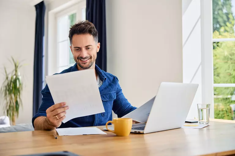 A man at a table looking at papers next to a laptop.