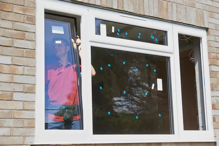 A man installing a large impact window onto a home with a tan brick exterior.