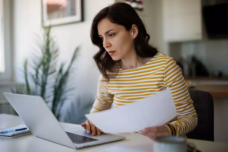 Woman holding documents with an open laptop.